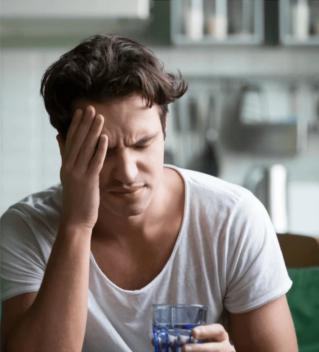 Man wincing with his hand on his forehead, holding a glass of water while hungover in his kitchen